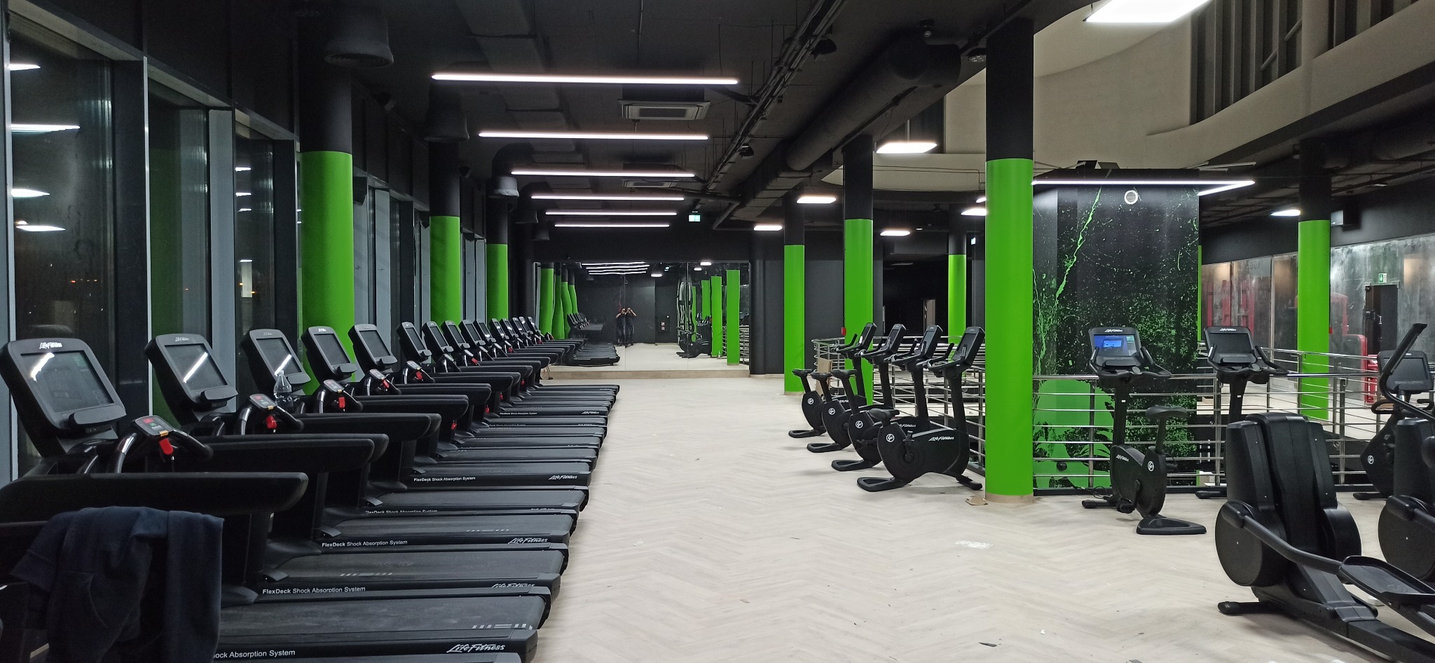 Nowoczesna siłownia z rzędem bieżni Life Fitness i rowerami stacjonarnymi, oświetlona liniowymi lampami LED, z zielonymi akcentami na kolumnach.