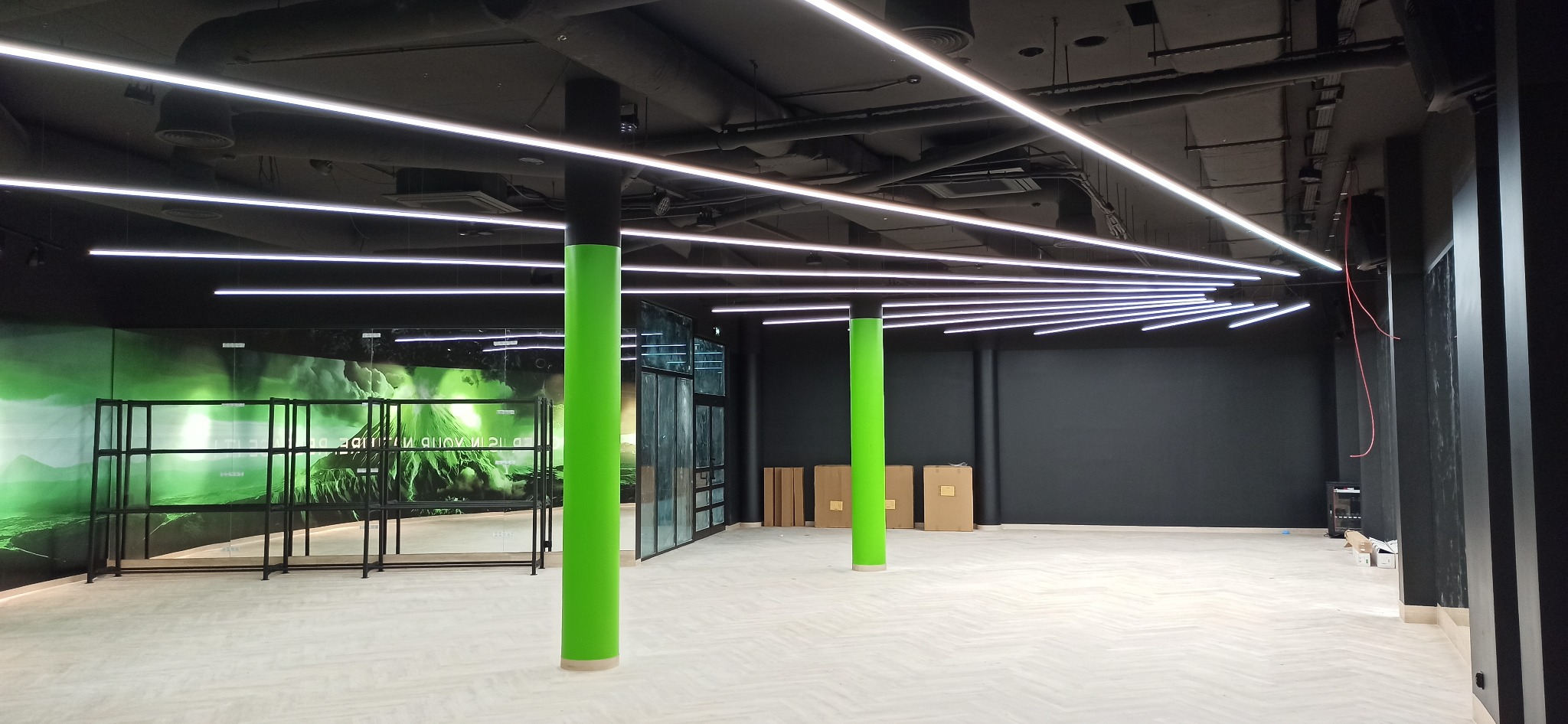 Nowoczesne oświetlenie LED w lokalu komercyjnym: równoległe, białe linie świetlne na czarnym suficie, zielone kolumny, surowy stan wykończenia.