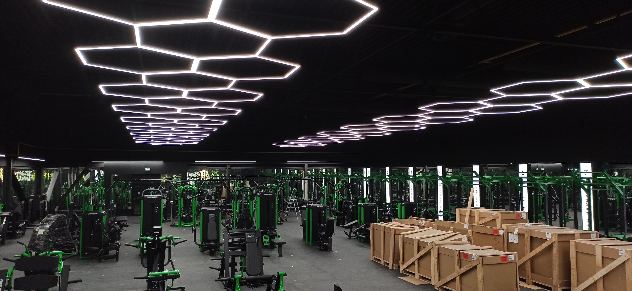 Nowoczesne oświetlenie LED w kształcie plastra miodu, zainstalowane w siłowni w Żołyni. Widoczne maszyny do ćwiczeń i drewniane skrzynie.