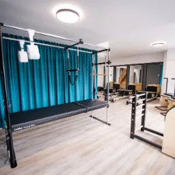 Jasne studio z profesjonalnym sprzętem do ćwiczeń: Cadillac Trapeze Table, Reformer i Ladder Barrel. Turkusowe zasłony w tle.