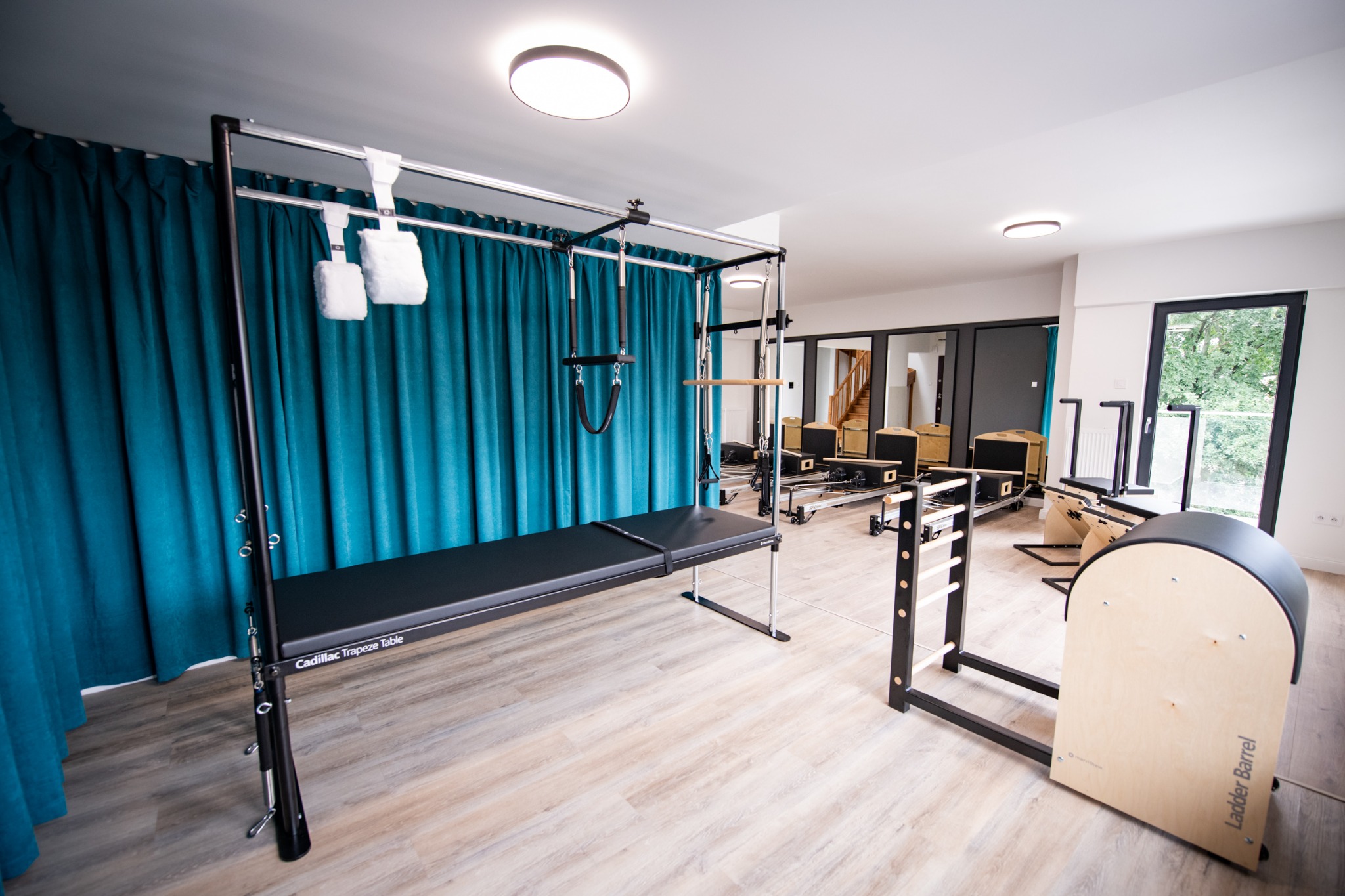 Jasne studio z profesjonalnym sprzętem do ćwiczeń: Cadillac Trapeze Table, Reformer i Ladder Barrel. Turkusowe zasłony w tle.