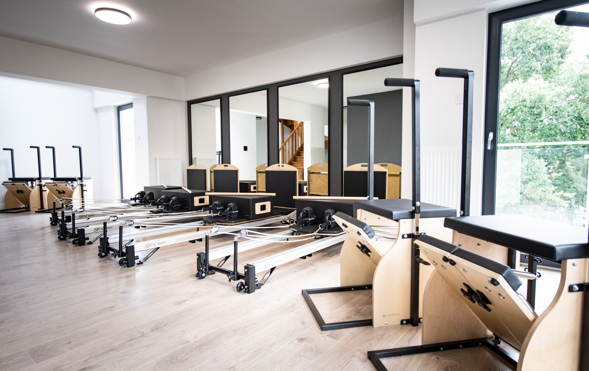 Jasne studio z rzędem reformerów do ćwiczeń, lustra na ścianie odbijają schody w tle.