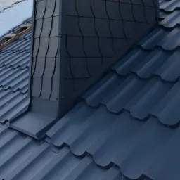 Nowoczesny dach z blachy w kolorze grafitowym sąsiadujący z kominem obłożonym łupkiem, widoczny fragment ośnieżonego dachu w tle.