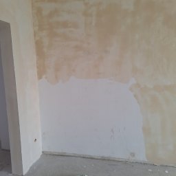 Usługi Budowlane - Przygotowanie ściany do malowania: fragment ściany z nałożoną warstwą gruntującą, widoczne niedoskonałości i plamy, na podłodze leży zielona szmatka w kratę.
