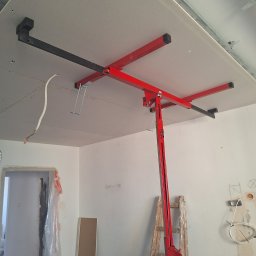 Usługi Budowlane - Czerwony podnośnik do płyt kartonowo-gipsowych podtrzymuje szarą płytę na suficie, widoczne metalowe profile konstrukcyjne sufitu, wiszące przewody elektryczne, drabina i ściana z notatkami ołówkiem.