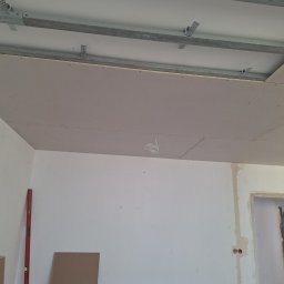 Usługi Budowlane - Montaż sufitu podwieszanego z płyt kartonowo-gipsowych na metalowej konstrukcji, widoczne profile i płyty przed szpachlowaniem, przewody elektryczne zwisające z sufitu, poziomica oparta o ścianę.