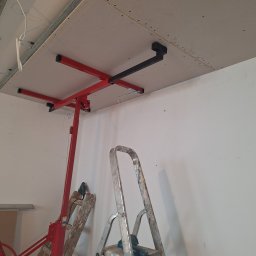 Usługi Budowlane - Montaż płyty gipsowo-kartonowej na suficie za pomocą podnośnika; widoczne profile metalowe, drabina i wkrętarka
