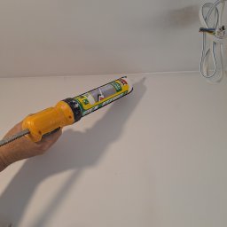 Usługi Budowlane - Aplikacja białego silikonu uszczelniającego z pistoletu na styku ściany i sufitu, widoczne wystające kable elektryczne.