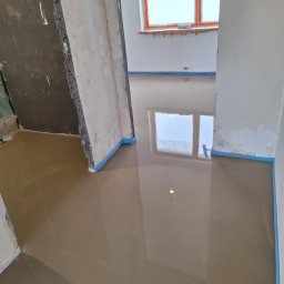 Usługi Budowlane - Wylewka samopoziomująca na podłodze w trakcie prac wykończeniowych, zabezpieczona niebieską taśmą malarską przy ścianach, widoczne surowe ściany i fragment okna.