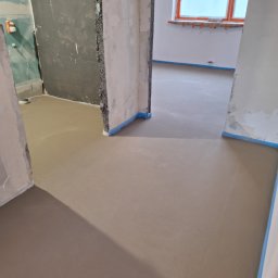 Usługi Budowlane - Wyrównana, świeża wylewka podłogowa w pomieszczeniu w trakcie remontu, zabezpieczona niebieską taśmą malarską przy ścianach i ościeżnicach, widoczne surowe ściany w tle.