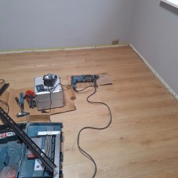 Usługi Budowlane - Przygotowane narzędzia do układania paneli podłogowych: wiertarka, poziomica, skrzynka z wiertłami, metalowa walizka na akumulatory, rękawice ochronne i młotek, na tle świeżo pomalowanej ściany...