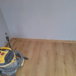 Fragment pokoju z nowo ułożoną podłogą z paneli, widoczny odkurzacz przemysłowy Stanley i listwy przypodłogowe z MDF w kolorze drewna.