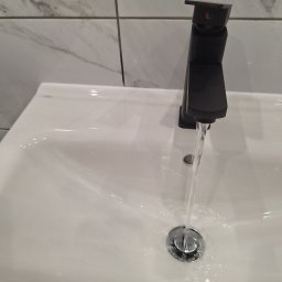 Usługi Budowlane - Czarna bateria umywalkowa z wypływającą wodą w Sękowo, na tle białych płytek imitujących marmur.
