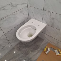 Usługi Budowlane - Wisząca, biała miska WC marki Koło, zamontowana na ścianie w łazience wyłożonej szarymi płytkami imitującymi marmur, z widocznymi opakowaniami po kleju na podłodze zabezpieczonej kartonem.