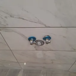 Białe płytki imitujące marmur z zamontowanymi zaworami kątowymi z niebieskimi osłonami, widoczne narzędzia i akcesoria montażowe na podłodze.