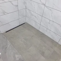 Wykończona podłoga i ściany w łazience wyłożone płytkami w odcieniach szarości i bieli, z widocznym odpływem liniowym.