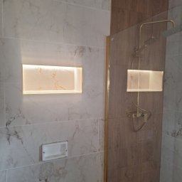 Usługi Budowlane - Wykończona łazienka z marmurowymi i drewnopodobnymi płytkami, oświetloną wnęką i złotą armaturą. Widoczny odpływ liniowy.