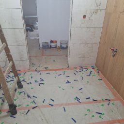 Usługi Budowlane - Fragment łazienki w trakcie prac glazurniczych: częściowo wyłożone płytki imitujące marmur, poziomowanie z użyciem klinów i krzyżyków, drabina. Widoczne narzędzia i materiały.
