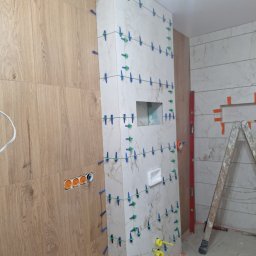Usługi Budowlane - Ściana w trakcie układania płytek imitujących drewno i marmur, z użyciem poziomicy i systemu poziomowania. Widoczne otwory pod instalacje i drabina.