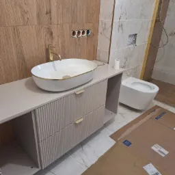 Nowoczesna łazienka po remoncie: biała umywalka nablatowa z złotą armaturą, szafka z ryflowanym frontem, podwieszana toaleta i marmurowe płytki.