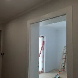 Wnętrze po malowaniu, widoczna biała rama drzwi, drabina oparta o ścianę, poziomica przy oknie zabezpieczonym folią malarską, resztki farby na podłodze.