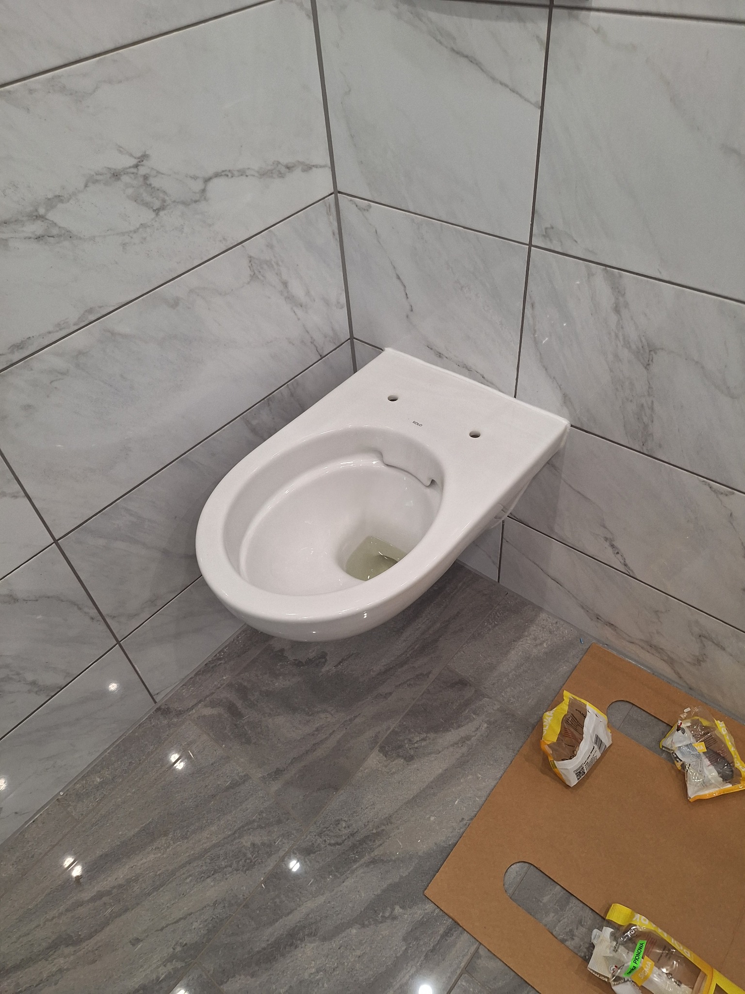 Wisząca, biała miska WC marki Koło, zamontowana na ścianie w łazience wyłożonej szarymi płytkami imitującymi marmur, z widocznymi opakowaniami po kleju na podłodze zabezpieczonej kartonem.