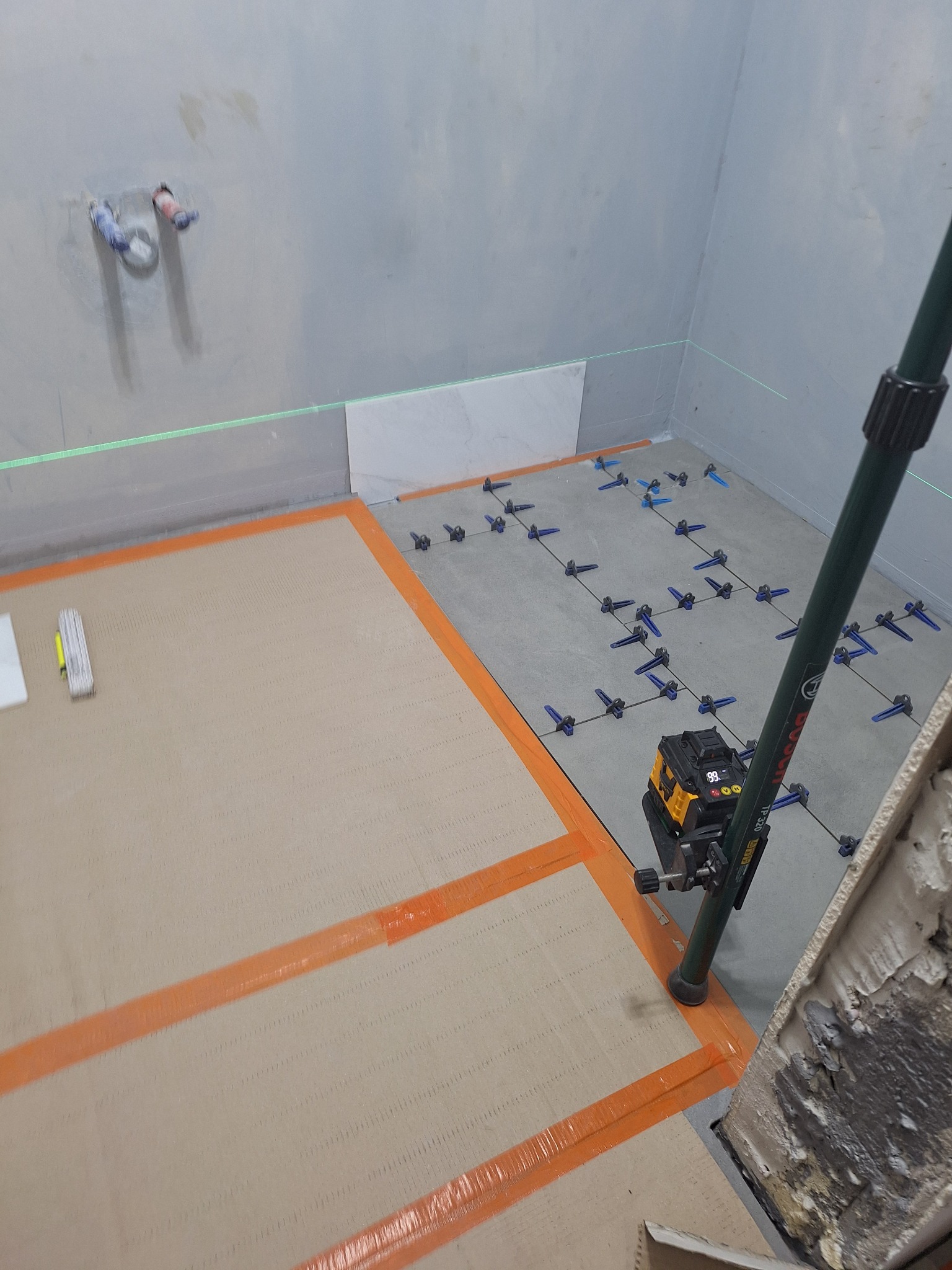 Przygotowanie do układania płytek w łazience, widoczny laser krzyżowy Bosch TP 320, poziomowanie podłoża, elementy dystansowe do płytek i częściowo ułożona pierwsza płytka na ścianie.