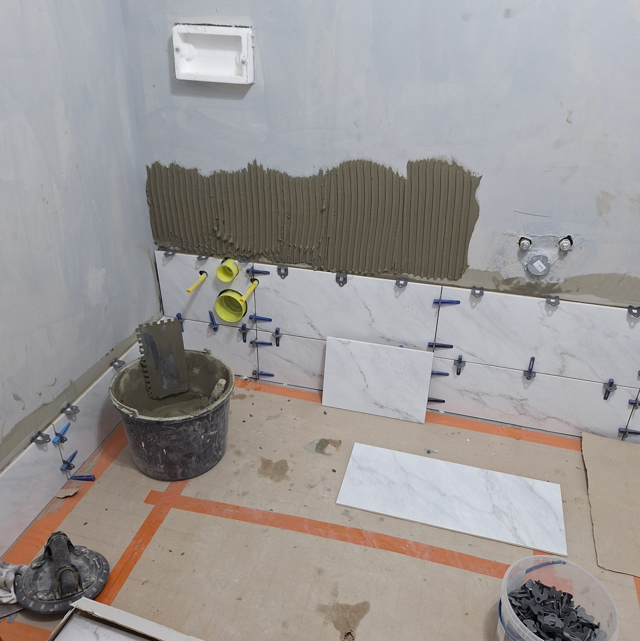 Ściana łazienki w trakcie układania płytek imitujących marmur, widoczne żółte rury, wiadro z zaprawą klejową, oraz system poziomowania płytek.
