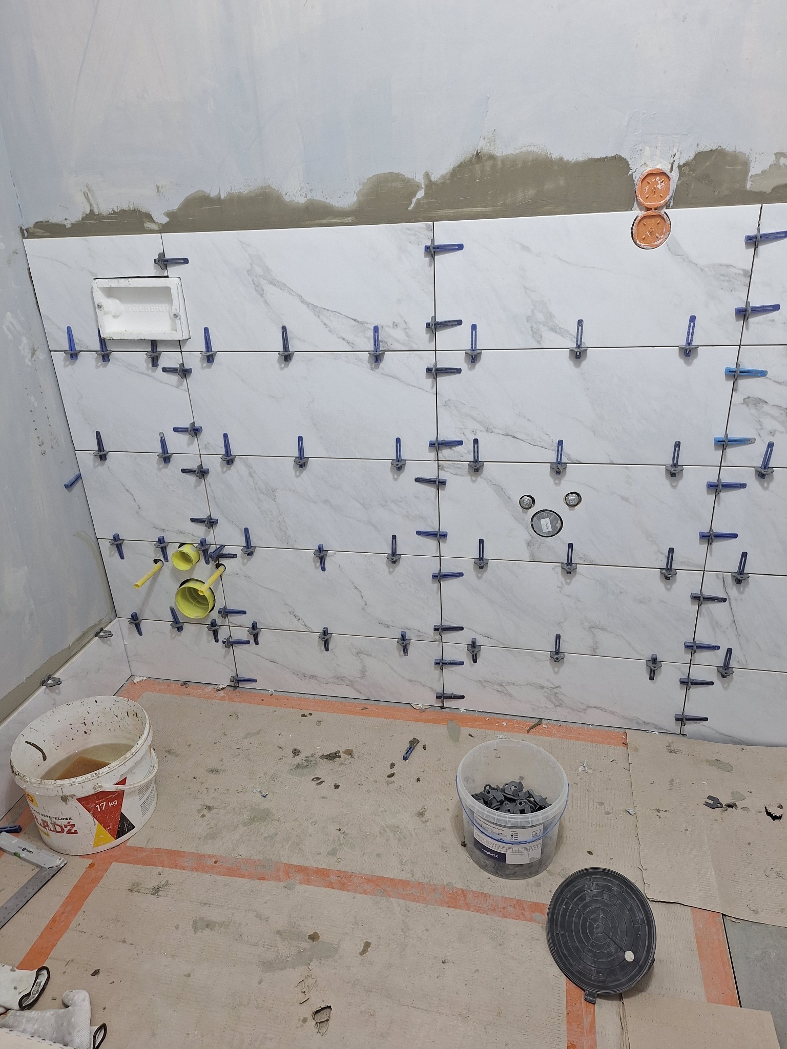 Ściana łazienki w trakcie układania płytek imitujących marmur, z użyciem klipsów poziomujących, widoczne przygotowane otwory na instalacje i sanitariaty, oraz dwa wiadra na podłodze.