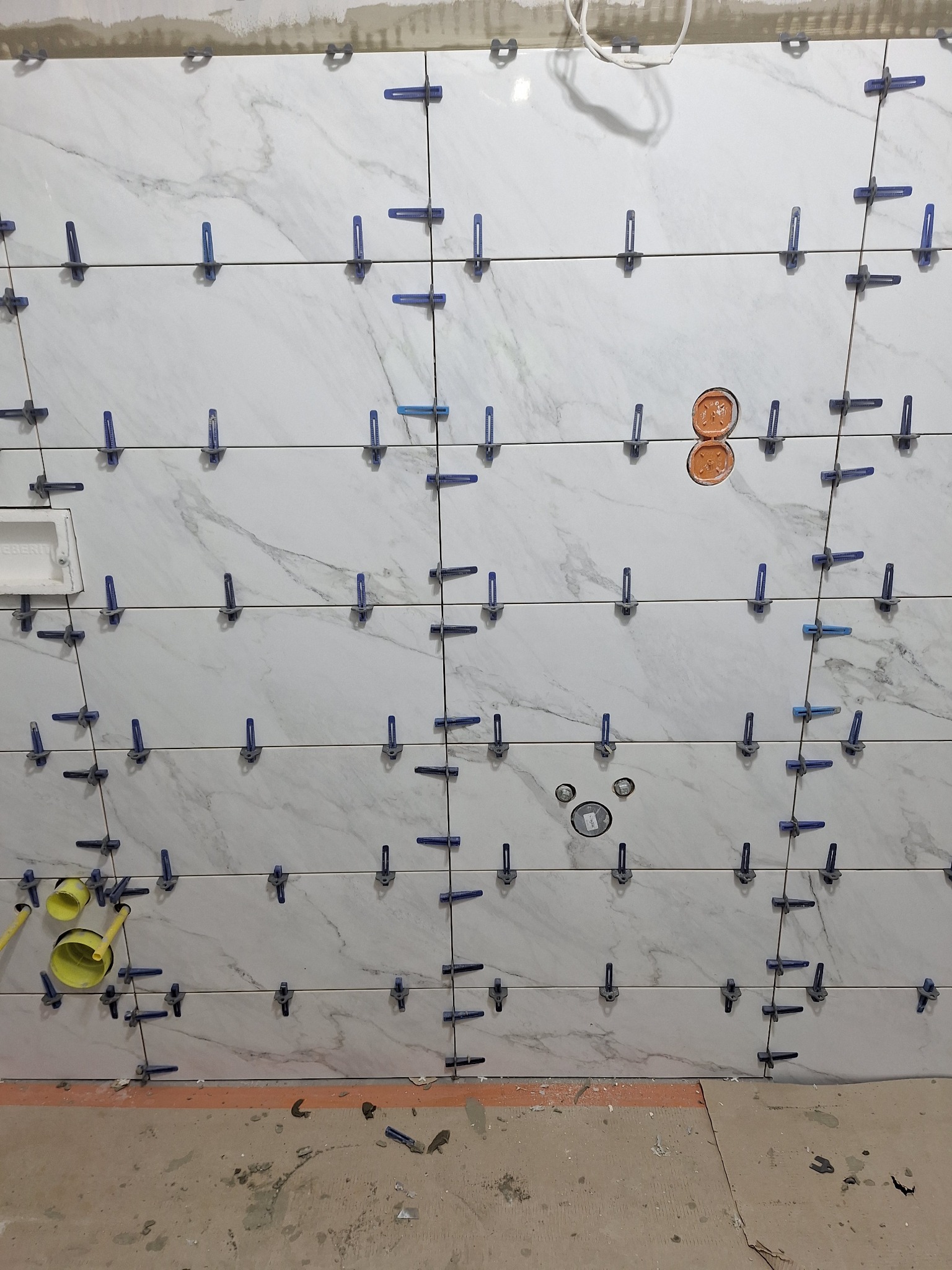 Ściana z białych płytek imitujących marmur, z niebieskimi klipsami poziomującymi, widoczne otwory na instalacje i fragment podłogi w trakcie prac wykończeniowych.