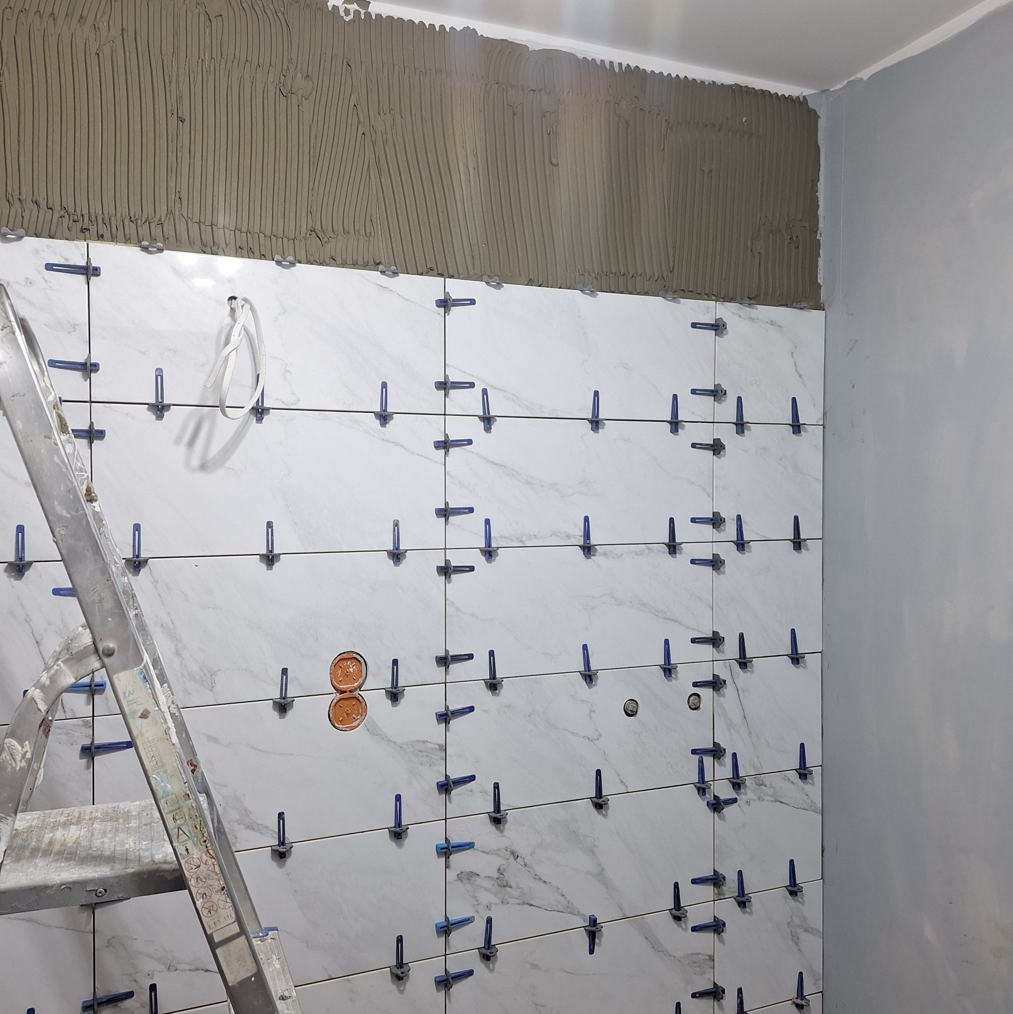 Ściana w trakcie układania marmuropodobnych płytek z użyciem niebieskich klipsów poziomujących; widoczna drabina malarska i przygotowana powierzchnia pod dalsze płytki.