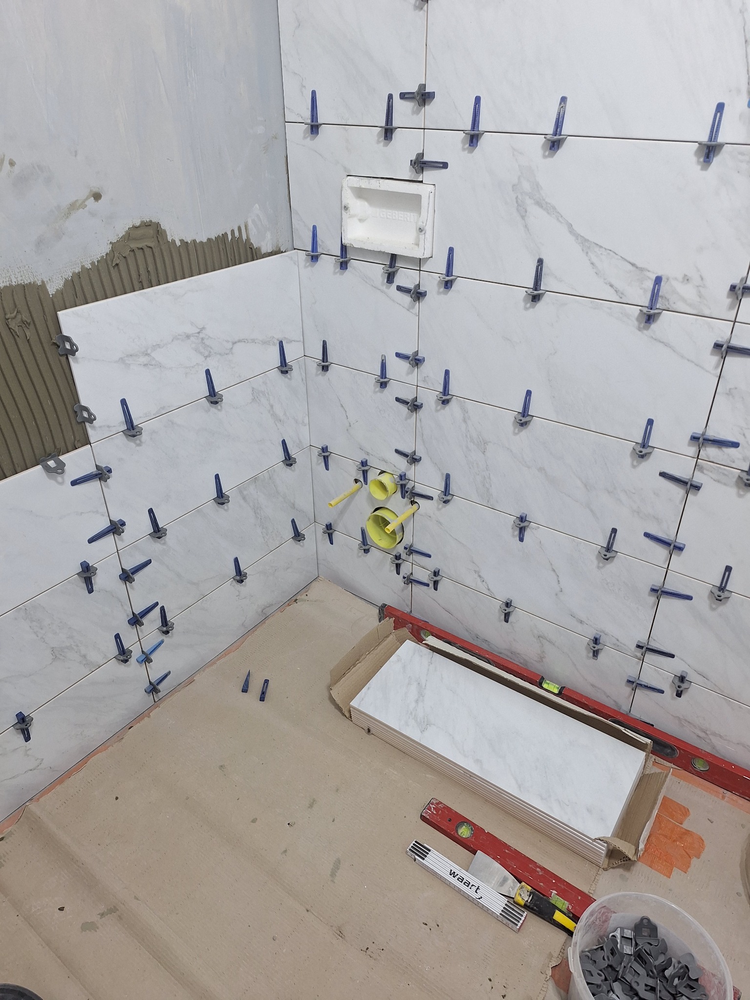 Ściana w trakcie układania płytek imitujących marmur, z użyciem niebieskich krzyżyków dystansowych, widoczne poziomice i puszka instalacyjna.