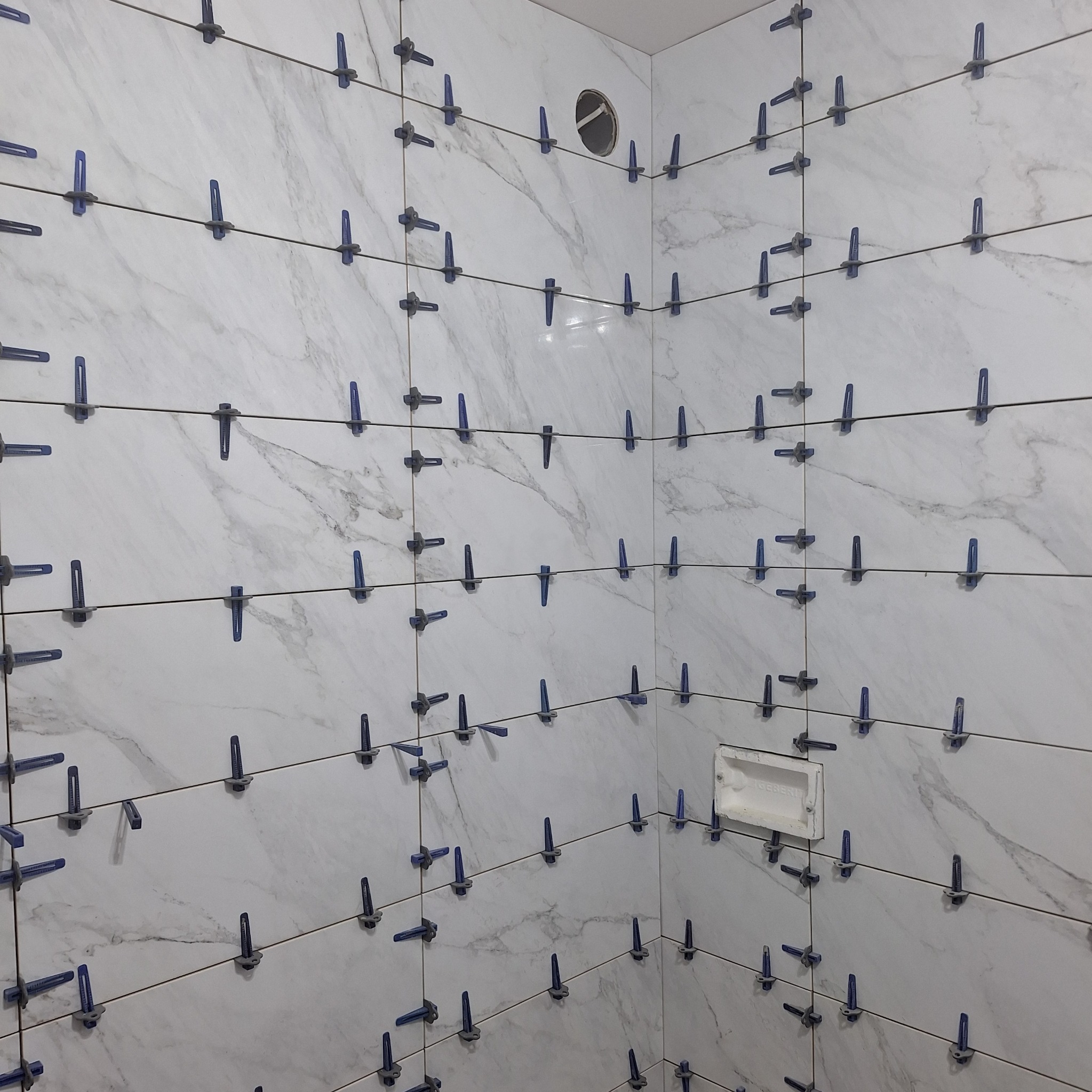 Świeżo ułożone, marmurowe płytki w rogu łazienki, z niebieskimi klipsami poziomującymi i sznurkiem, widoczna puszka instalacyjna podtynkowego elementu.