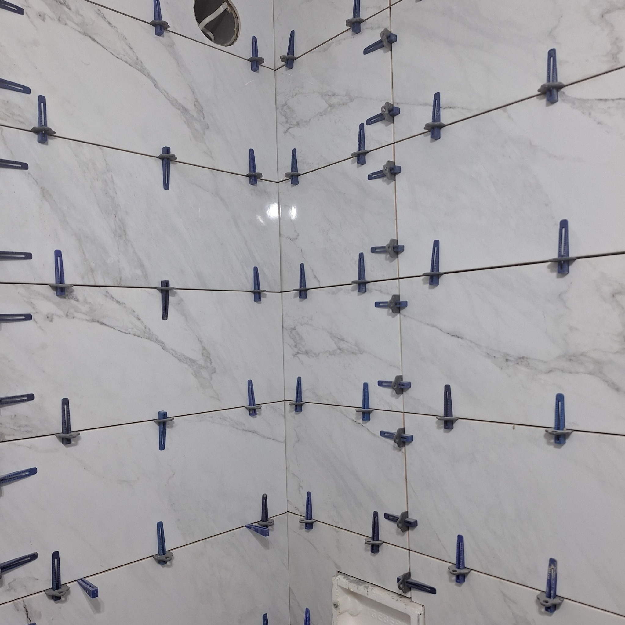 Ściana wyłożona białymi płytkami imitującymi marmur, z niebieskimi krzyżykami dystansowymi, widoczne przewody wystające z otworu w górnej części ściany.