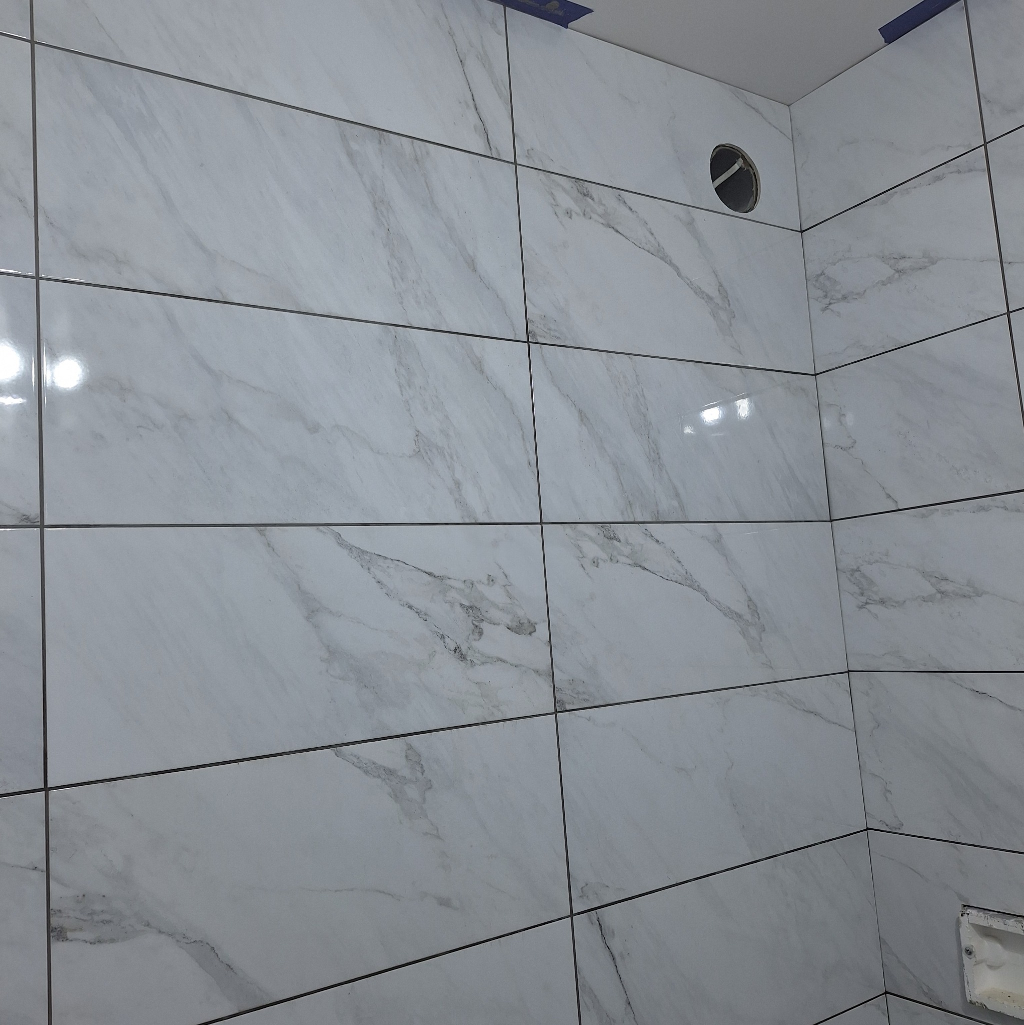 Wykończona ściana łazienki z białych, marmurkowych płytek ceramicznych ułożonych poziomo, widoczny otwór na rurę i fragment instalacji elektrycznej w dolnym rogu.