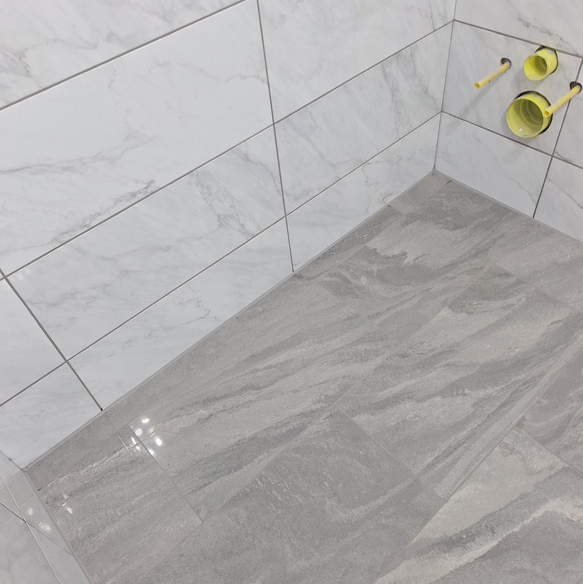 Wykończona narożna przestrzeń łazienki z szarymi płytkami podłogowymi imitującymi kamień i białymi marmurkowymi płytkami ściennymi, widoczne żółte rury instalacji wodnej.