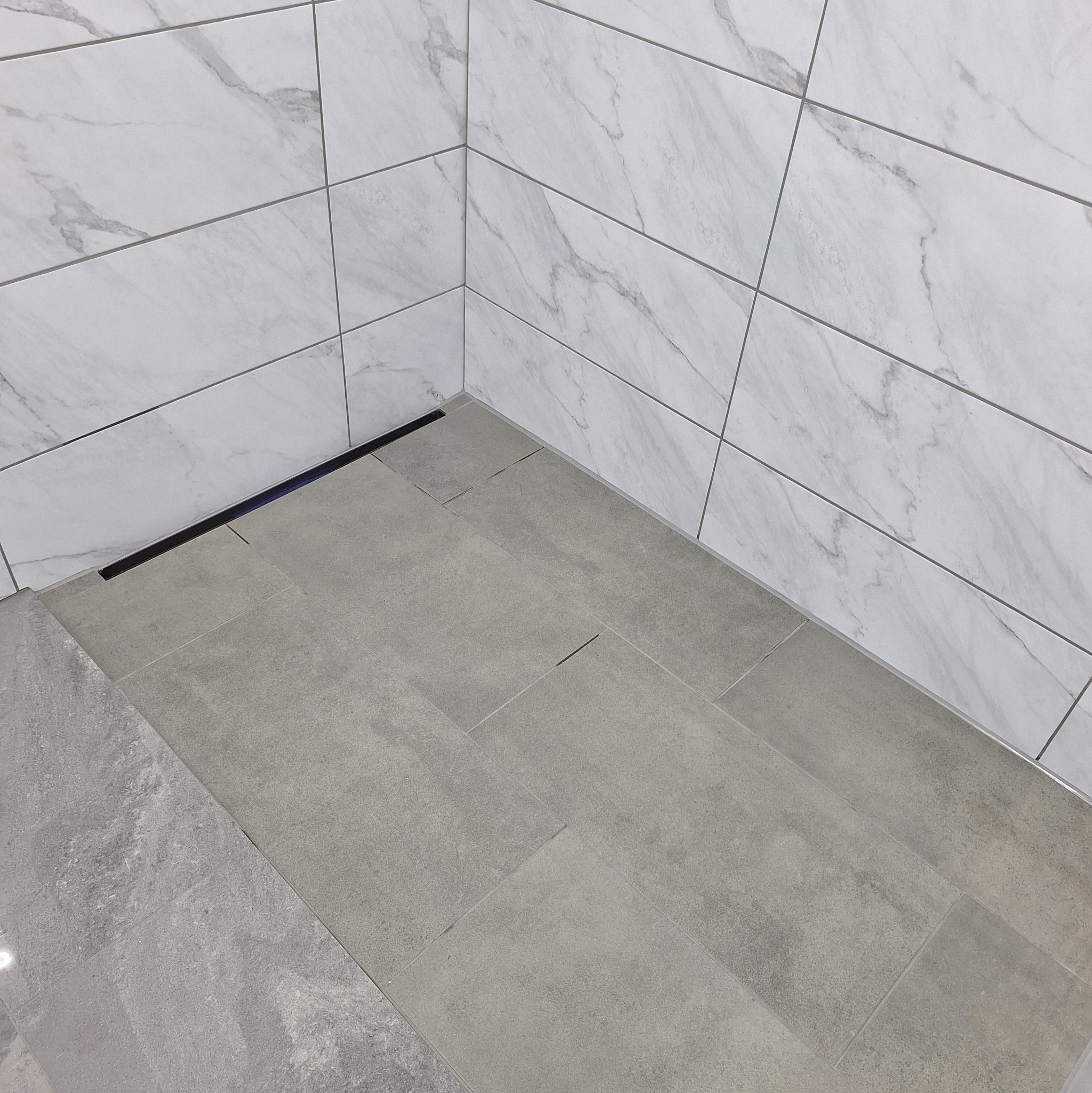 Wykończona podłoga i ściany w łazience wyłożone płytkami w odcieniach szarości i bieli, z widocznym odpływem liniowym.