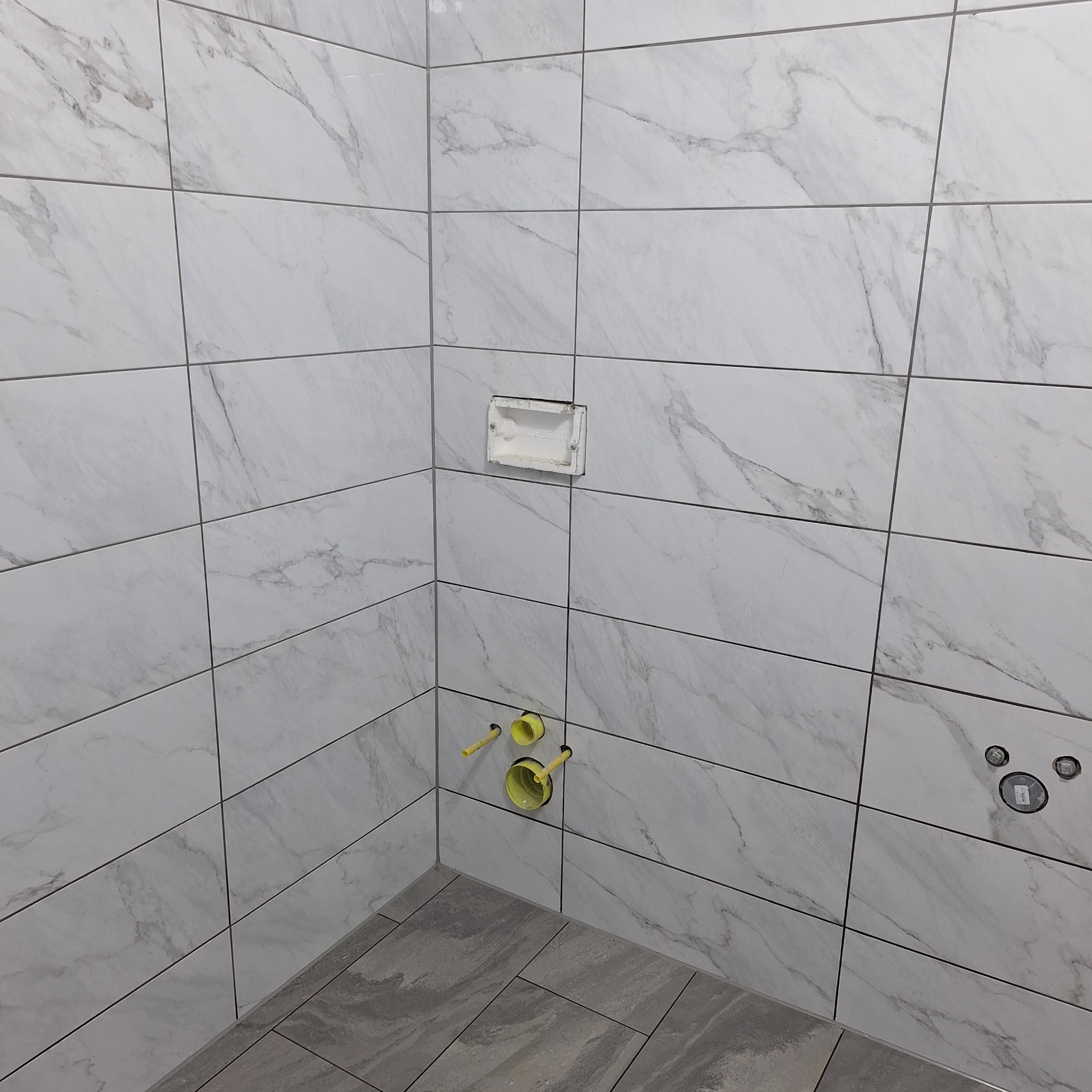 Wykończona ściana łazienki z białymi płytkami imitującymi marmur, widoczne podłączenia hydrauliczne i otwór na zabudowę podtynkową toalety.