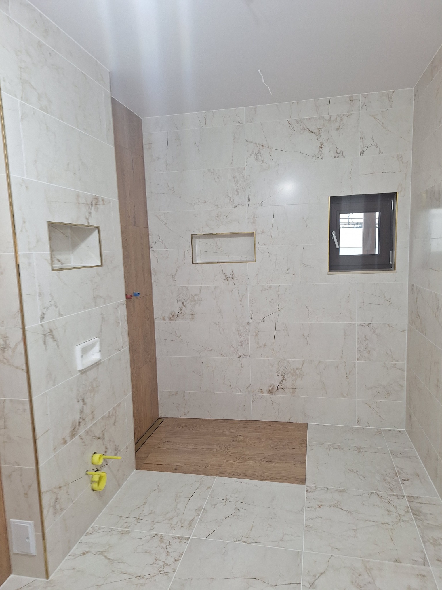 Wykończona łazienka z marmurowymi płytkami, drewnianą podłogą w kabinie prysznicowej i wnękami w ścianach. Widoczne przyłącza wodne i okno.