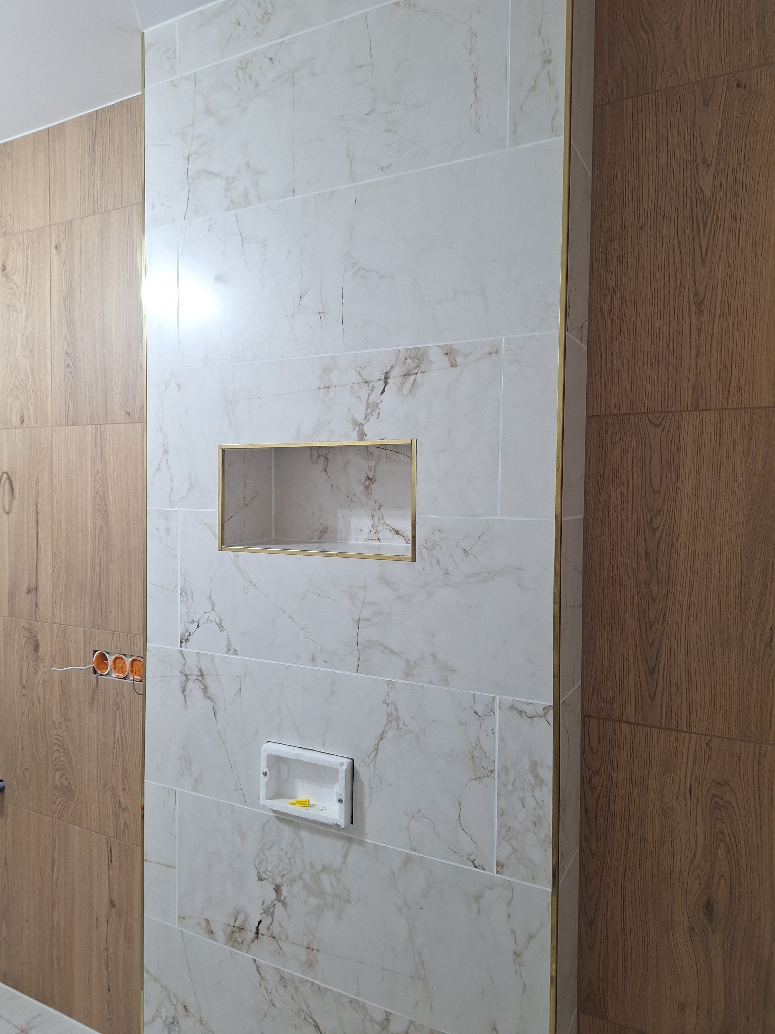 Ściana wykończona płytkami imitującymi marmur z wnękami. Widoczne gniazdko elektryczne i fragment ściany wyłożonej panelami drewnopodobnymi.