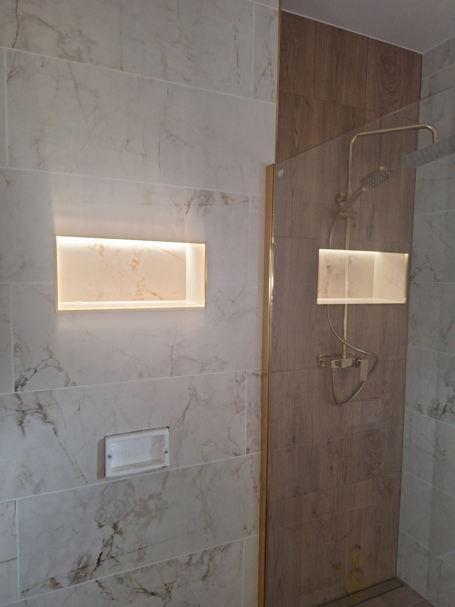 Wykończona łazienka z marmurowymi i drewnopodobnymi płytkami, oświetloną wnęką i złotą armaturą. Widoczny odpływ liniowy.