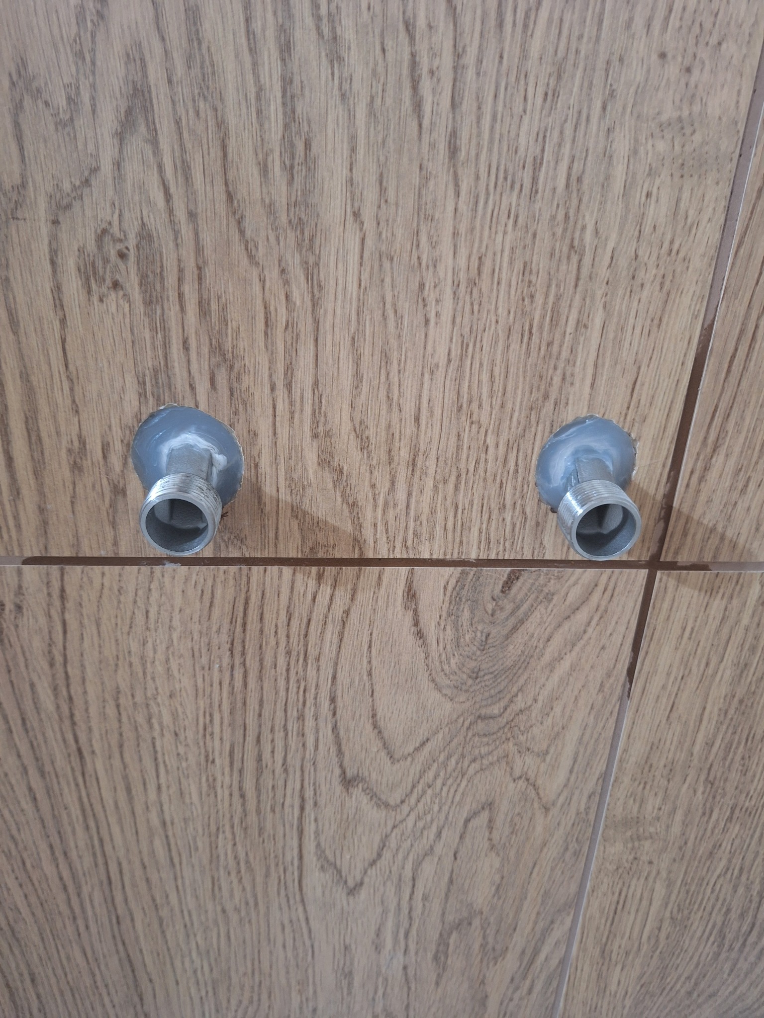 Ułożone płytki imitujące drewno z wystającymi przyłączami wodnymi, zabezpieczonymi silikonem. Widoczne fugi i faktura płytek. Detal wykończenia łazienki.