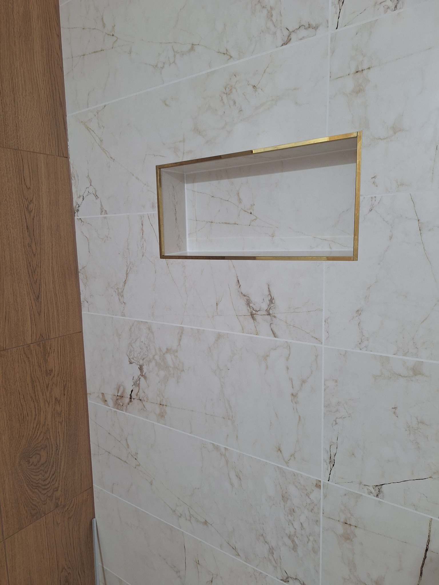 Wykończona ściana z marmurowymi płytkami i wnęką z obramowaniem w kolorze złota. Fragment drewnopodobnej okładziny po lewej stronie.