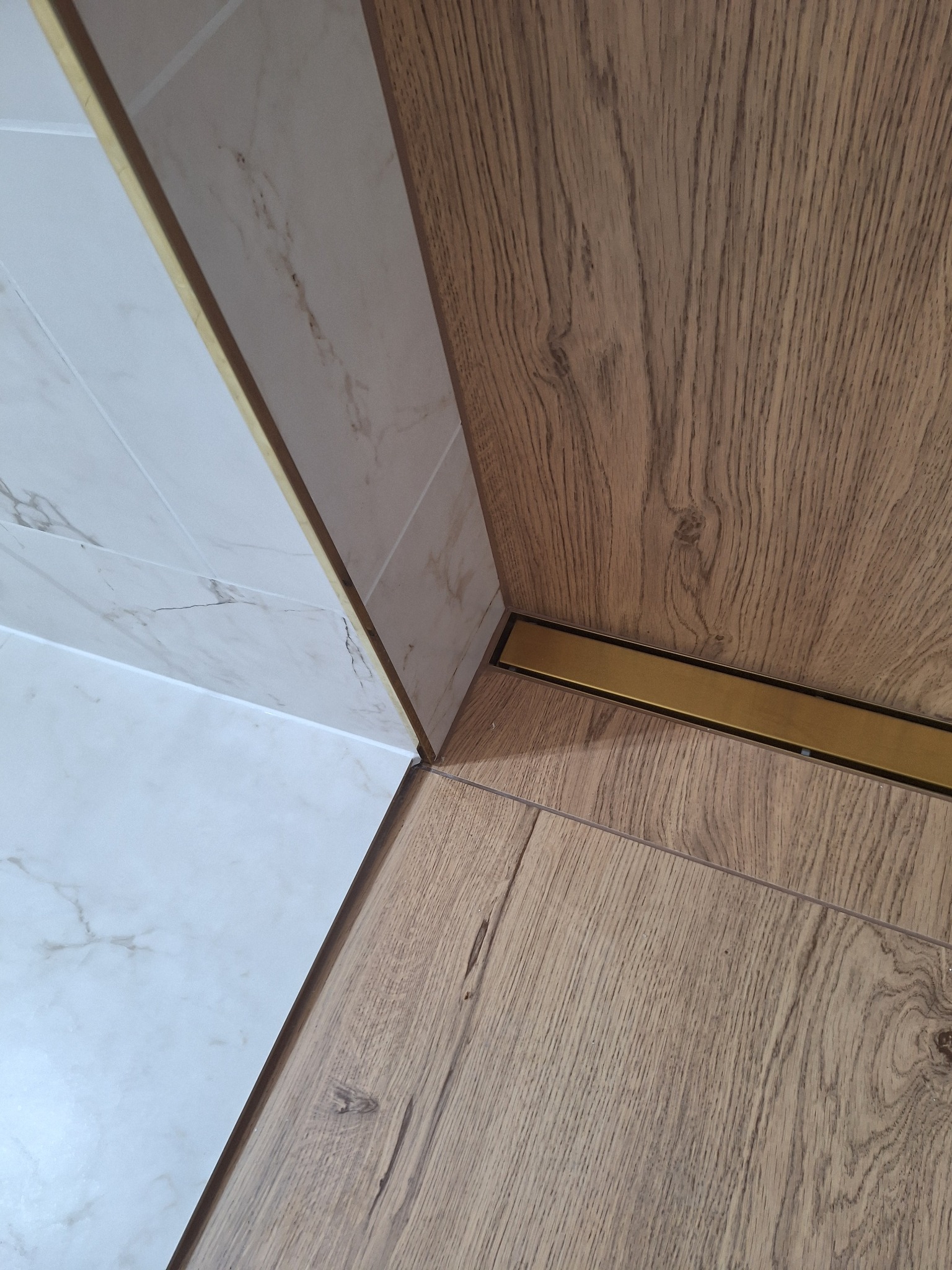 Połączenie płytek marmurowych z panelami podłogowymi, wykończone złotą listwą. Widoczne detale wykończenia podłogi i ściany w łazience.