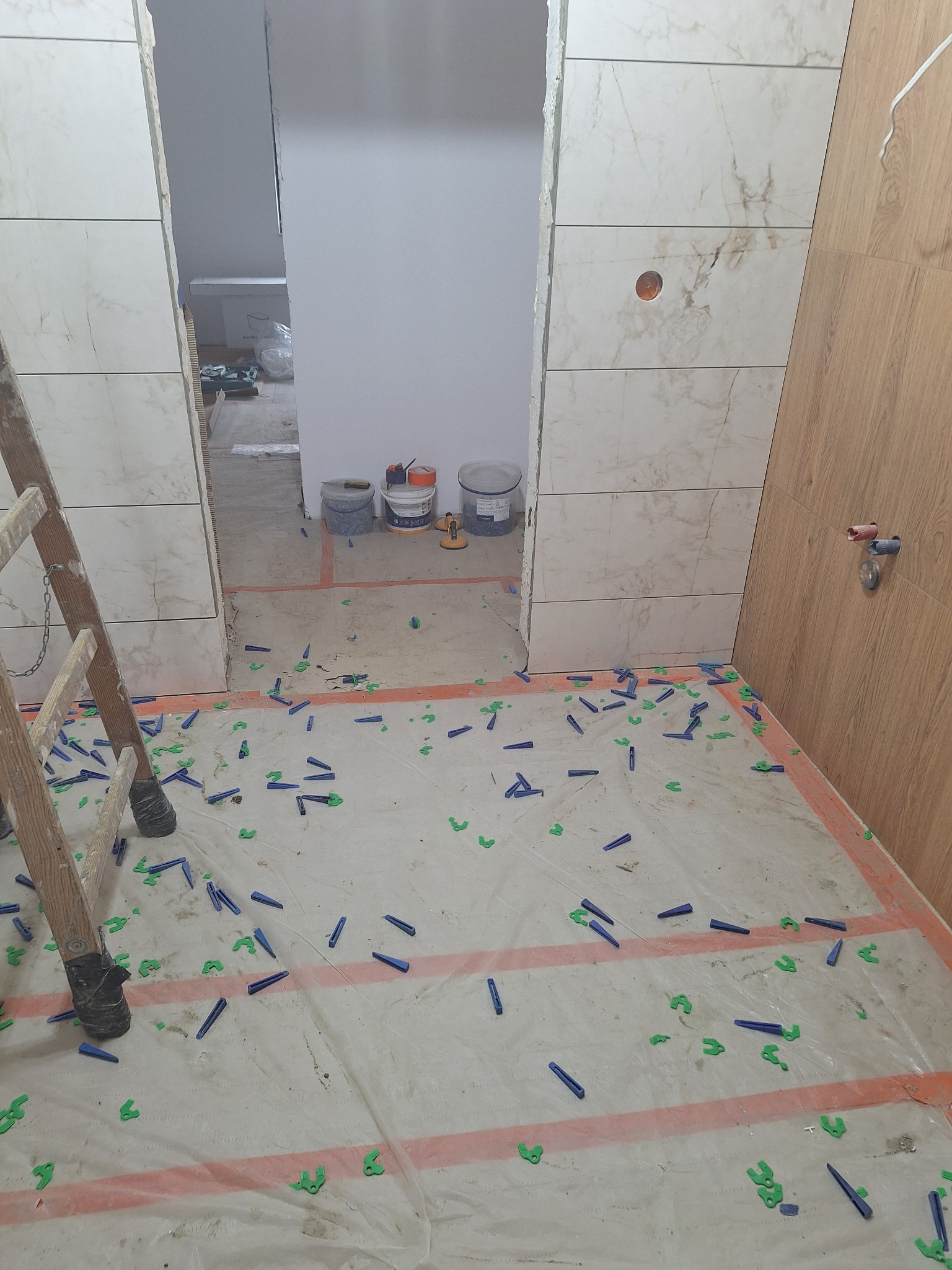 Fragment łazienki w trakcie prac glazurniczych: częściowo wyłożone płytki imitujące marmur, poziomowanie z użyciem klinów i krzyżyków, drabina. Widoczne narzędzia i materiały.