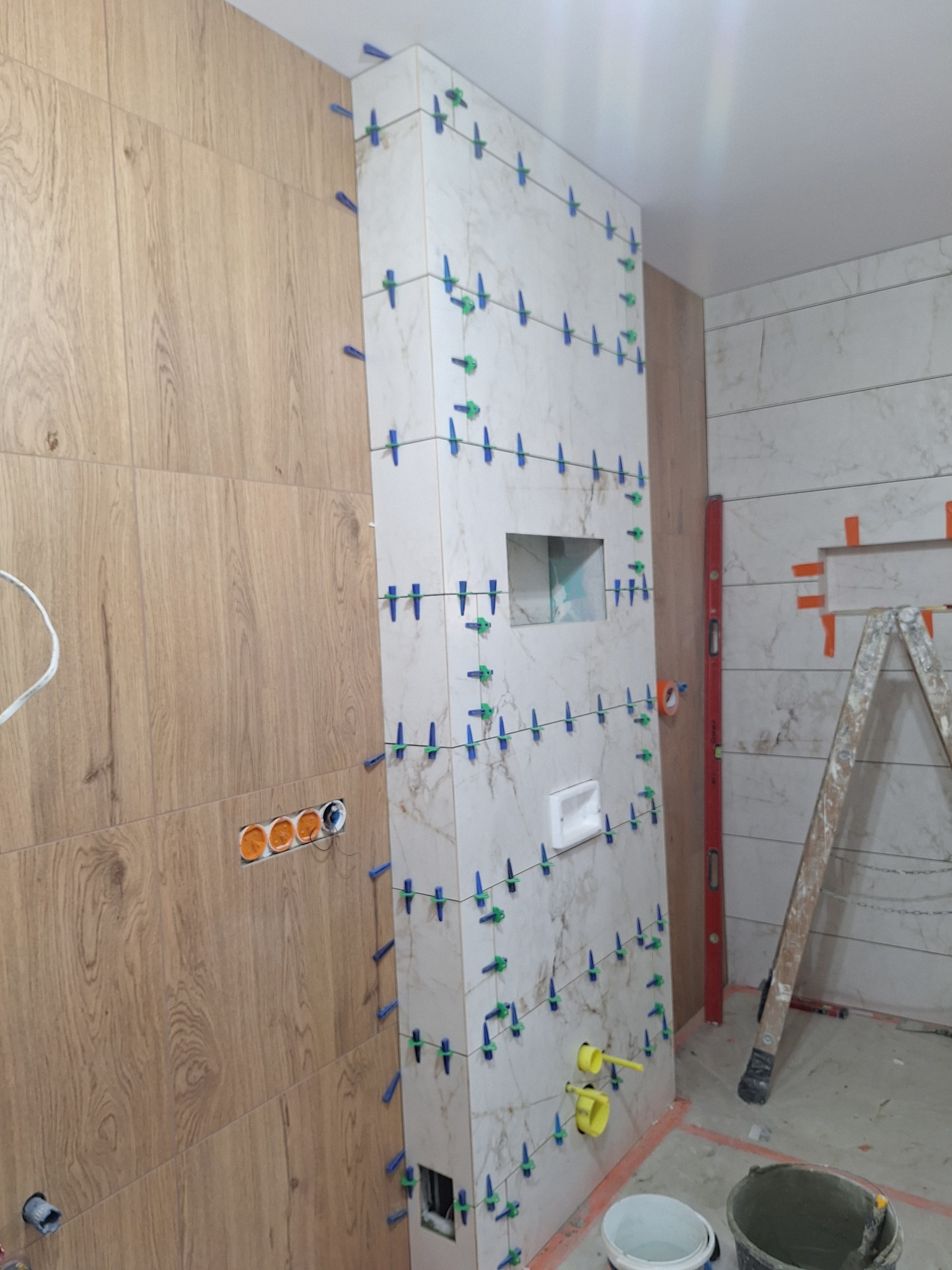 Ściana w trakcie układania płytek imitujących drewno i marmur, z użyciem poziomicy i systemu poziomowania. Widoczne otwory pod instalacje i drabina.