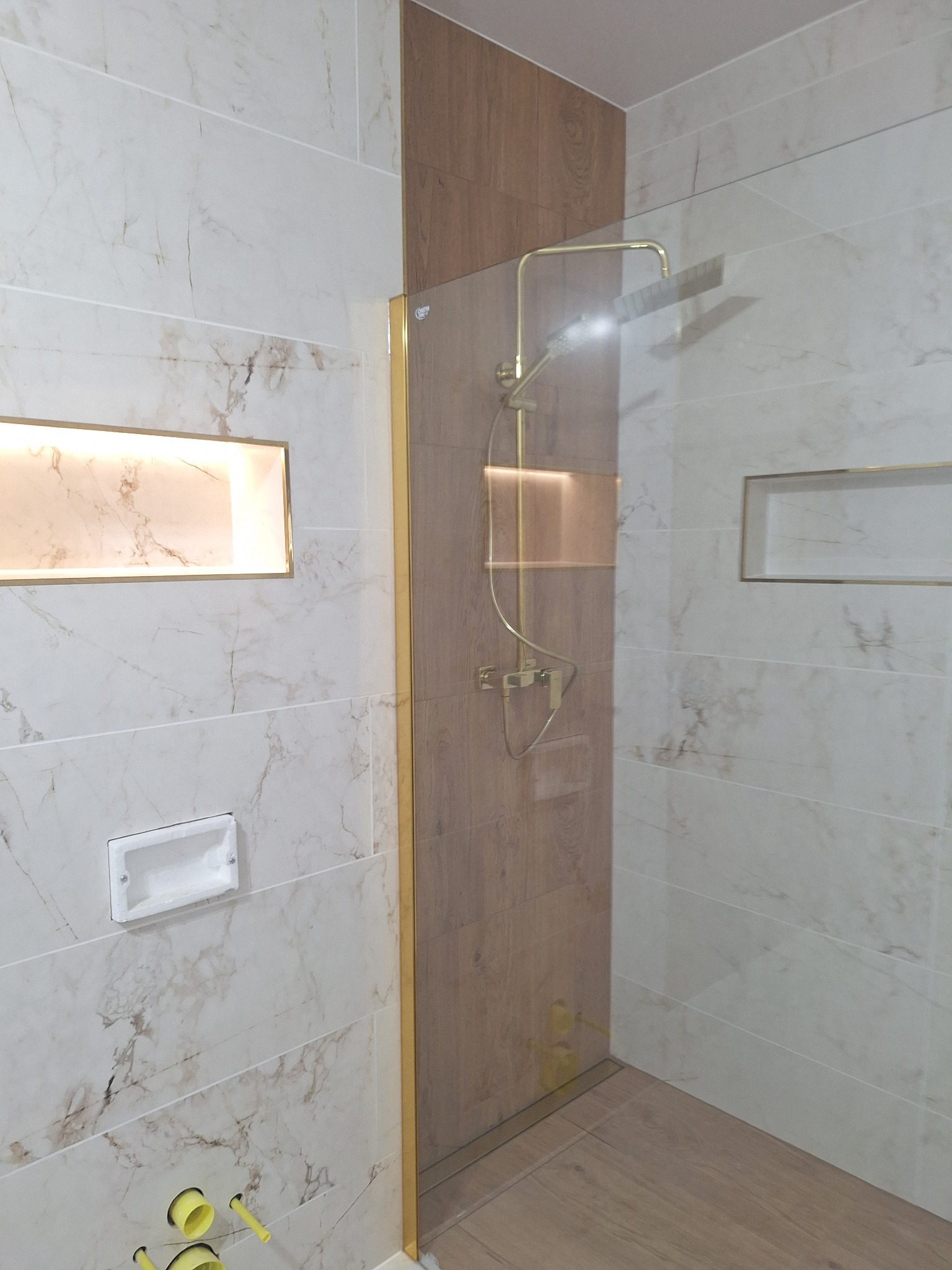 Nowoczesna łazienka z kabiną prysznicową typu walk-in, wykończona płytkami imitującymi marmur i drewno, ze złotymi akcentami i podświetlanymi niszami.