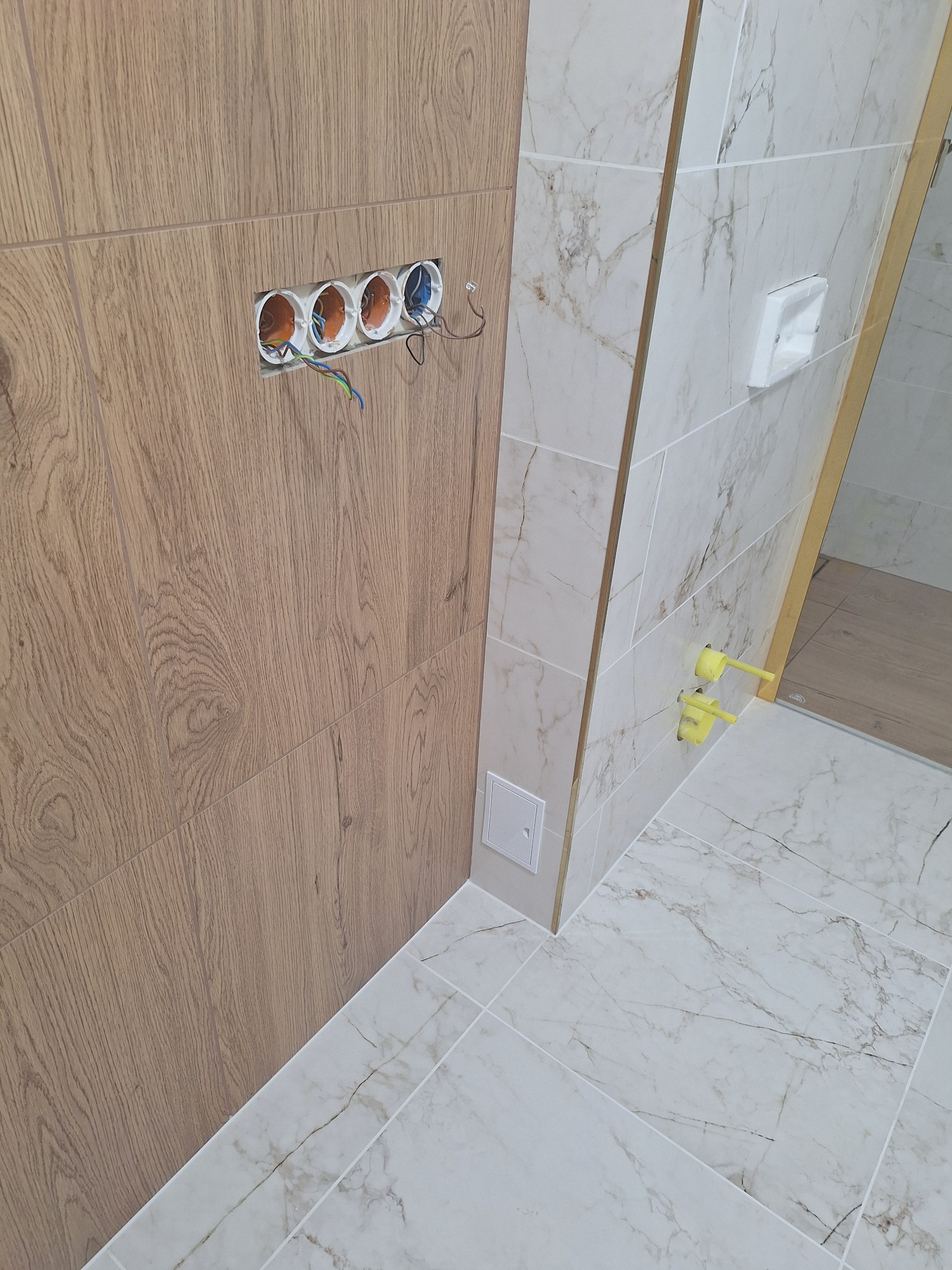 Wykończona ściana w łazience z drewnopodobnymi płytkami. Widoczne gniazdka elektryczne w trakcie montażu i fragment ściany z marmurowymi płytkami.