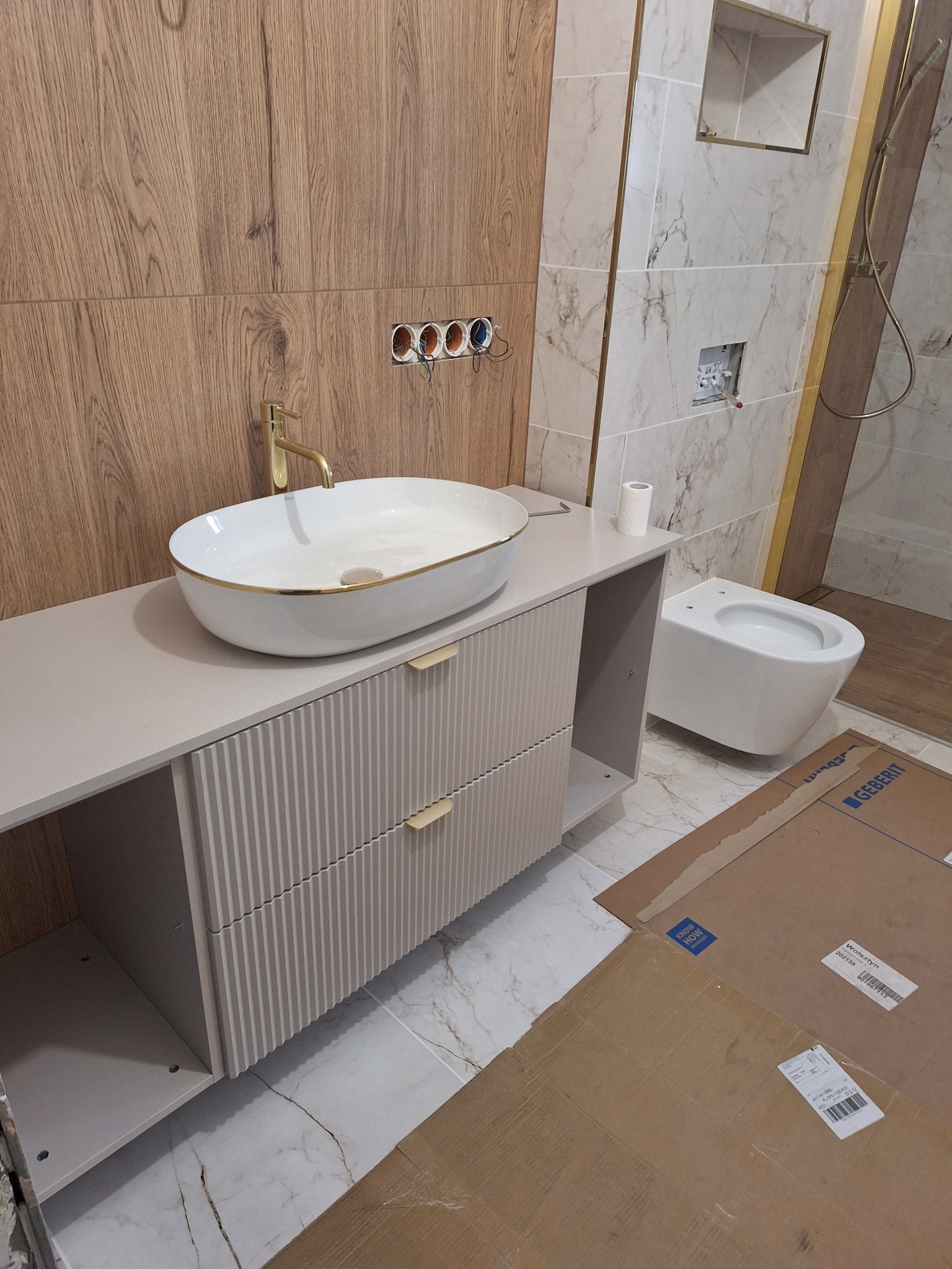 Nowoczesna łazienka po remoncie: biała umywalka nablatowa z złotą armaturą, szafka z ryflowanym frontem, podwieszana toaleta i marmurowe płytki.