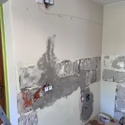 Ściana w trakcie szpachlowania i przygotowania do położenia płytek, widoczne gniazdka elektryczne, fragment instalacji wodnej i ślady po usuniętych płytkach.